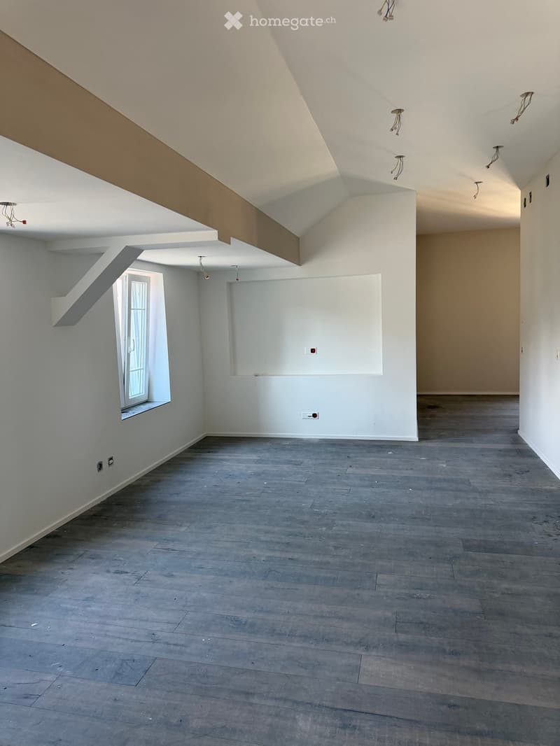 Empty room with grey wooden flooring, white walls, and a large window with a white frame. Exposed electrical wiring and light fixtures are visible on the ceiling.
