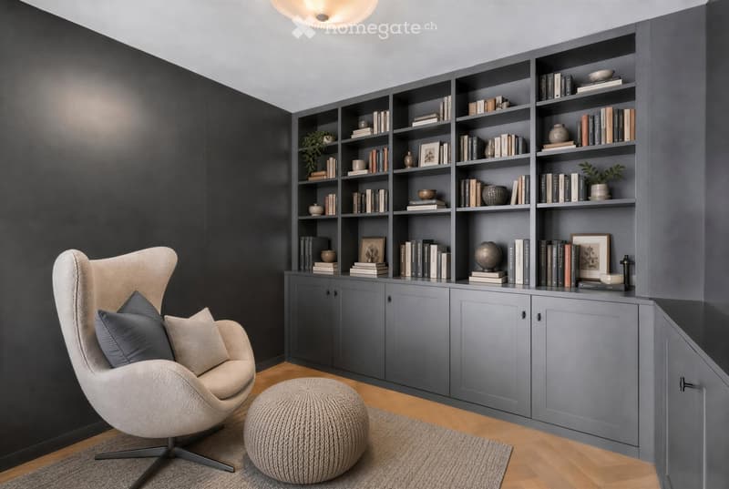 Modern living room with a grey bookshelf filled with books and decorative items, a beige armchair with pillows, and a round ottoman on a grey rug.