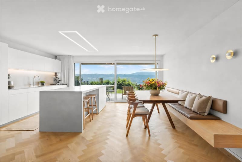 Modern kitchen with white cabinets, a central island, and a dining area with a wooden table and seating. Large windows offer a scenic view.