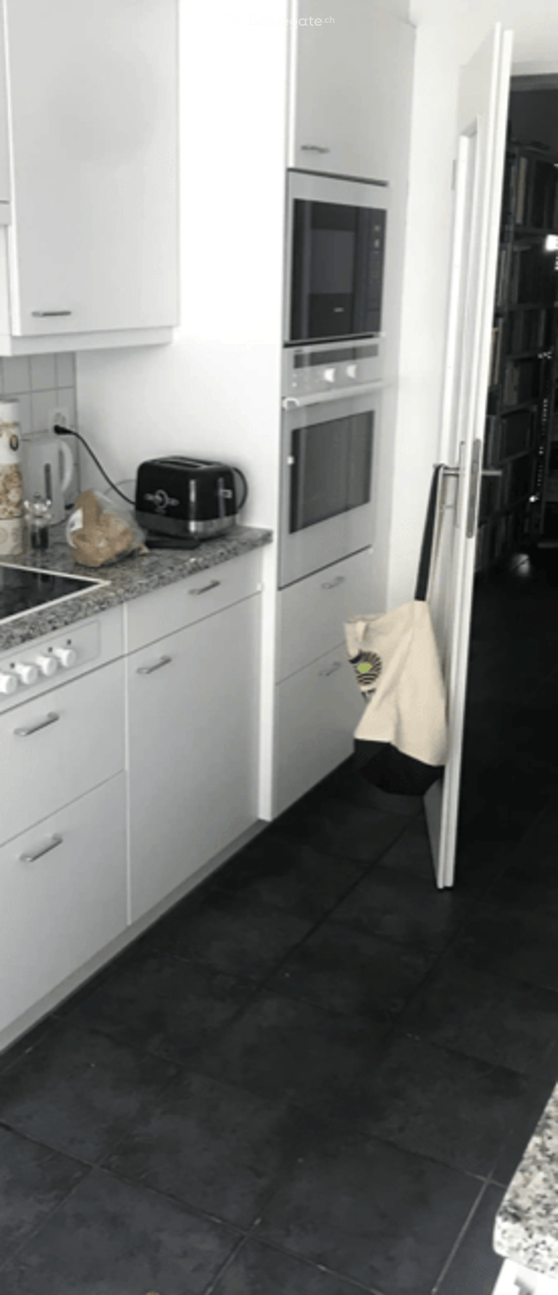 Modern kitchen with white cabinets, black tiled floor, microwave, oven, blender, toaster, and a handbag hanging on the door.