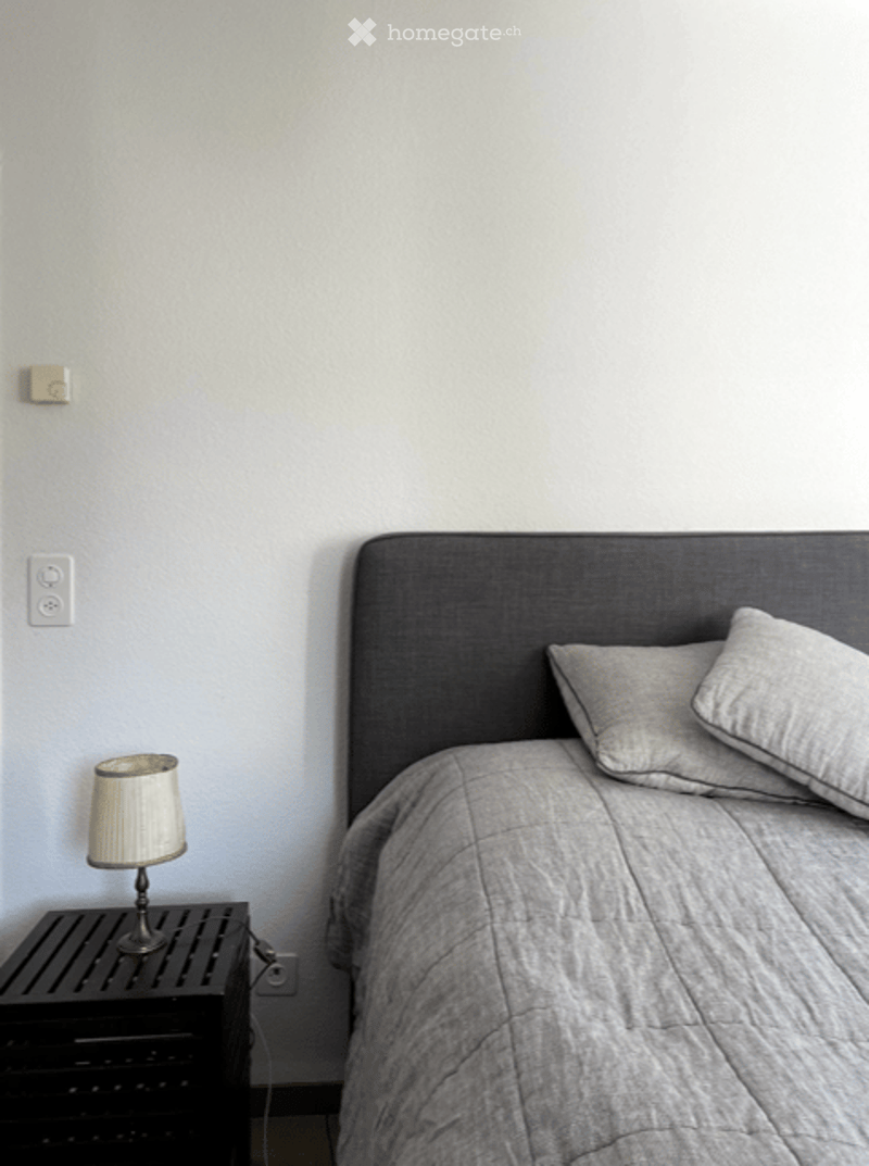 Minimalist bedroom with a grey headboard, a nightstand with a lamp, and a neatly made bed with pillows and a duvet.