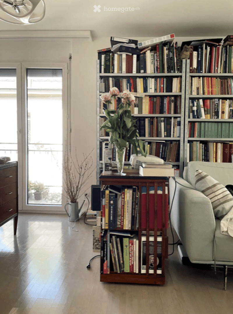 Living room with a light green sofa, wooden bookshelf filled with books, a small table with a vase of flowers, and a potted plant near the window.