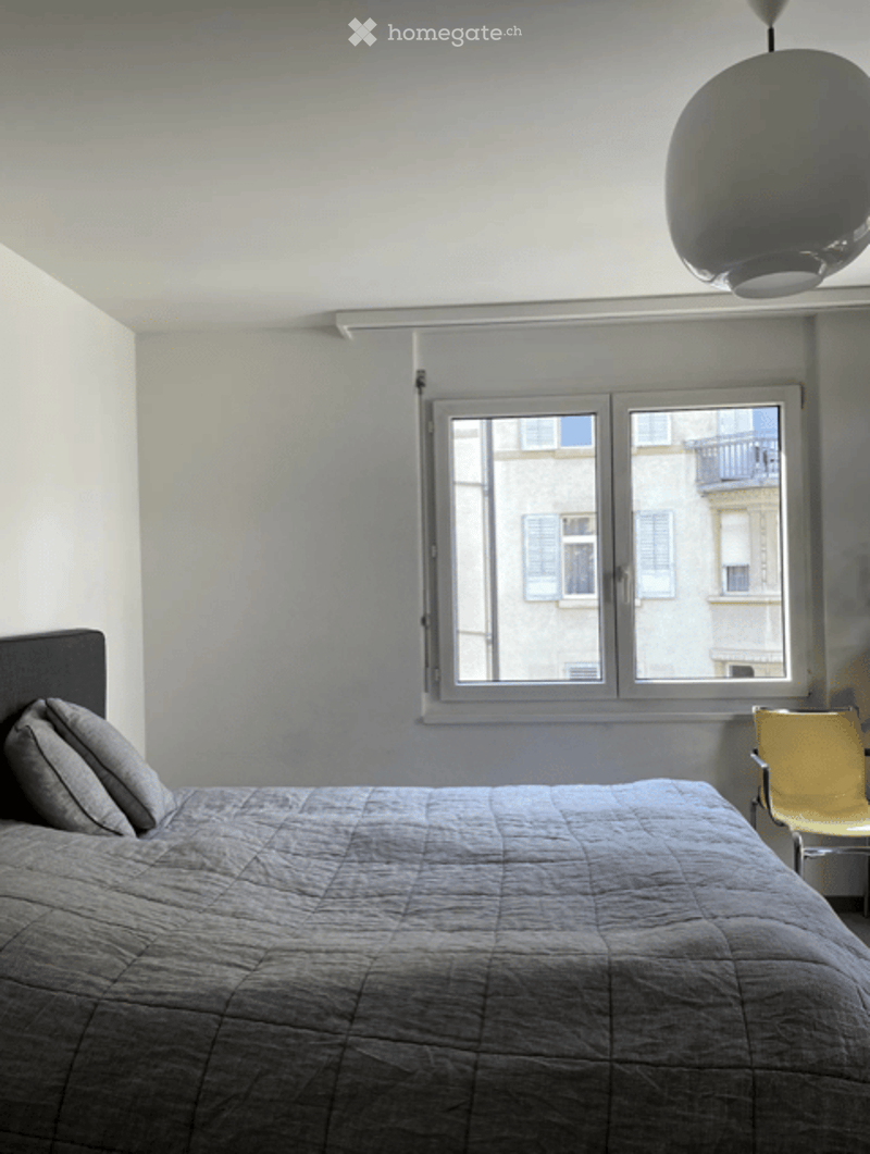 Bedroom with a grey bed, pillows, a yellow chair, and a large window with a view of a balcony.