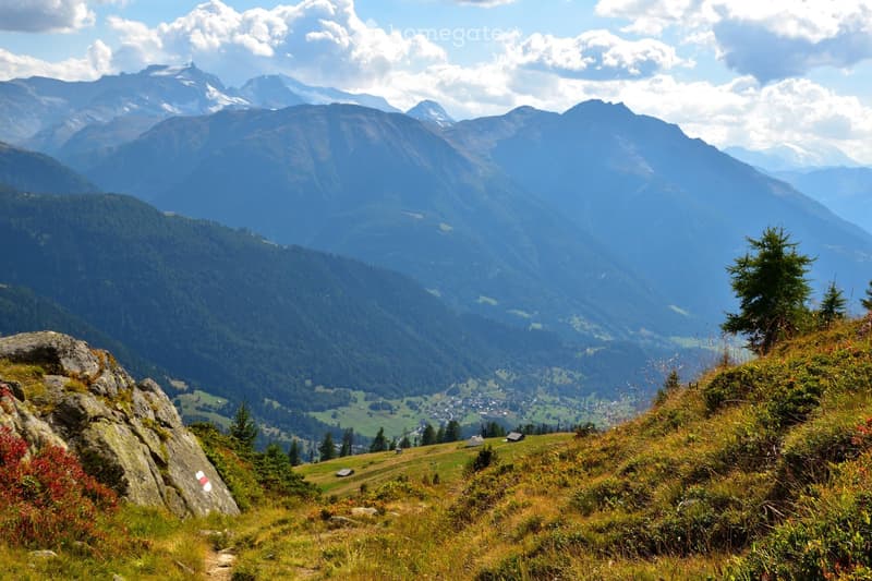 Panoramic mountain view with a valley below, scattered houses, and a winding path leading down the slope.
