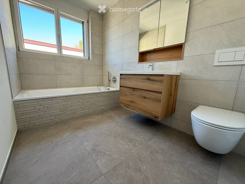 Modern bathroom with a bathtub, wooden sink cabinet, toilet, and a large mirror above the sink. The room features tiled walls and a tiled floor.