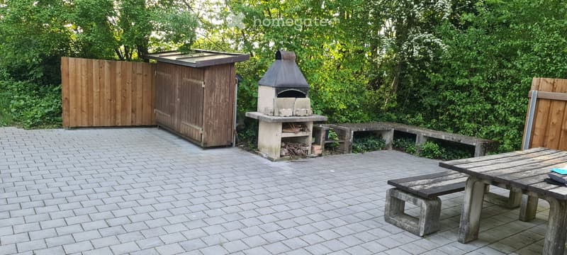 Outdoor patio area with brick flooring, wooden shed, stone barbecue, wooden table, and benches surrounded by greenery.