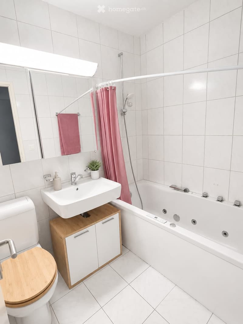 Modern bathroom with white tiles, a wooden toilet seat, a white sink with a wooden cabinet, a bathtub with a shower curtain, and a small potted plant on the sink.