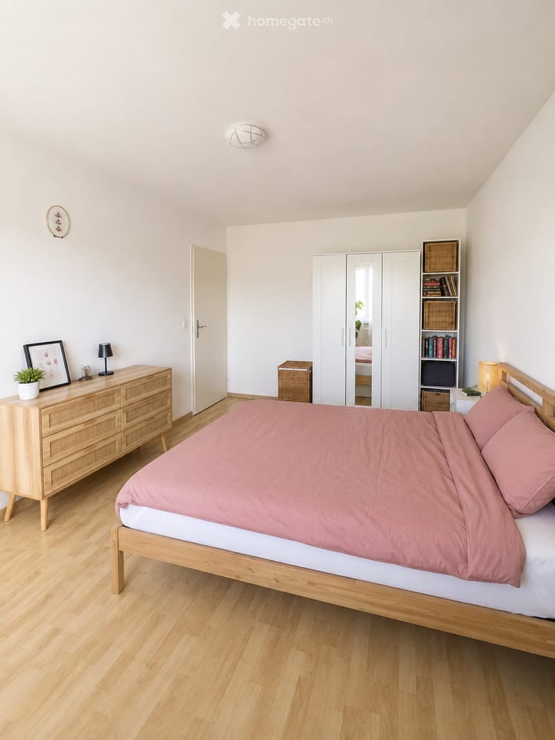 Bright bedroom with wooden flooring, a bed with pink bedding, a wooden nightstand with a lamp, a dresser with a picture frame and potted plant, a large mirrored wardrobe, and a bookshelf with books and baskets.