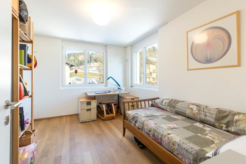 A small, well-lit bedroom with a wooden floor, a sofa bed, a desk with a chair, a shelf, and a framed picture on the wall. The room has large windows offering a view of the outdoors.