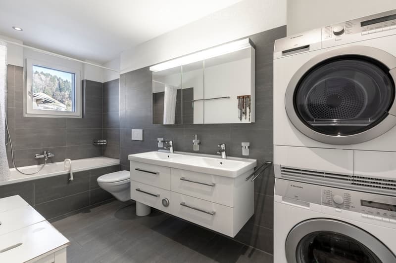 Modern bathroom with grey tiles, a bathtub, toilet, double sink, mirror, and a stacked washer/dryer unit.