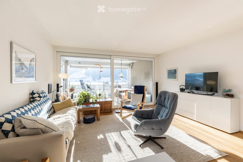 Bright living room with large windows and sliding glass doors leading to a balcony. Features a patterned sofa, coffee table, armchair, TV, floor lamp, and potted plants.