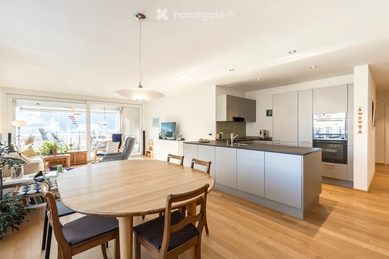 Open-plan living and dining area with a round wooden dining table, chairs, sofa, kitchen with cabinets, sink, and oven. Large windows offer a view of the balcony.