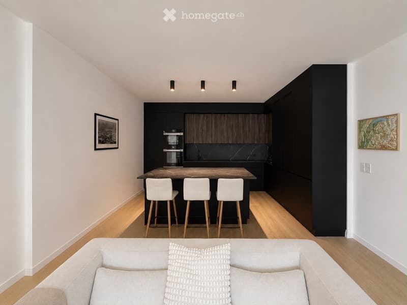 Modern living and dining area with a grey sofa, kitchen island, three white stools, dark cabinets, and artwork on the walls.
