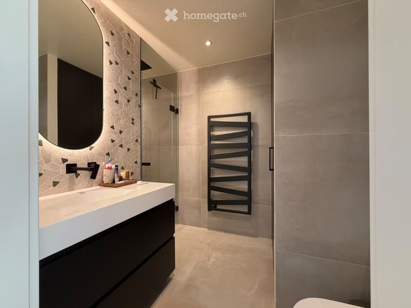 Modern bathroom with a white countertop sink, black faucet, large round mirror, and a glass-enclosed shower with a black towel rack.