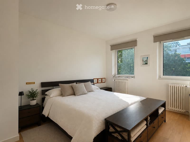 A well-lit bedroom with a large bed, nightstands, a dresser, and a potted plant. The room features wooden flooring, white walls, and large windows with blinds.