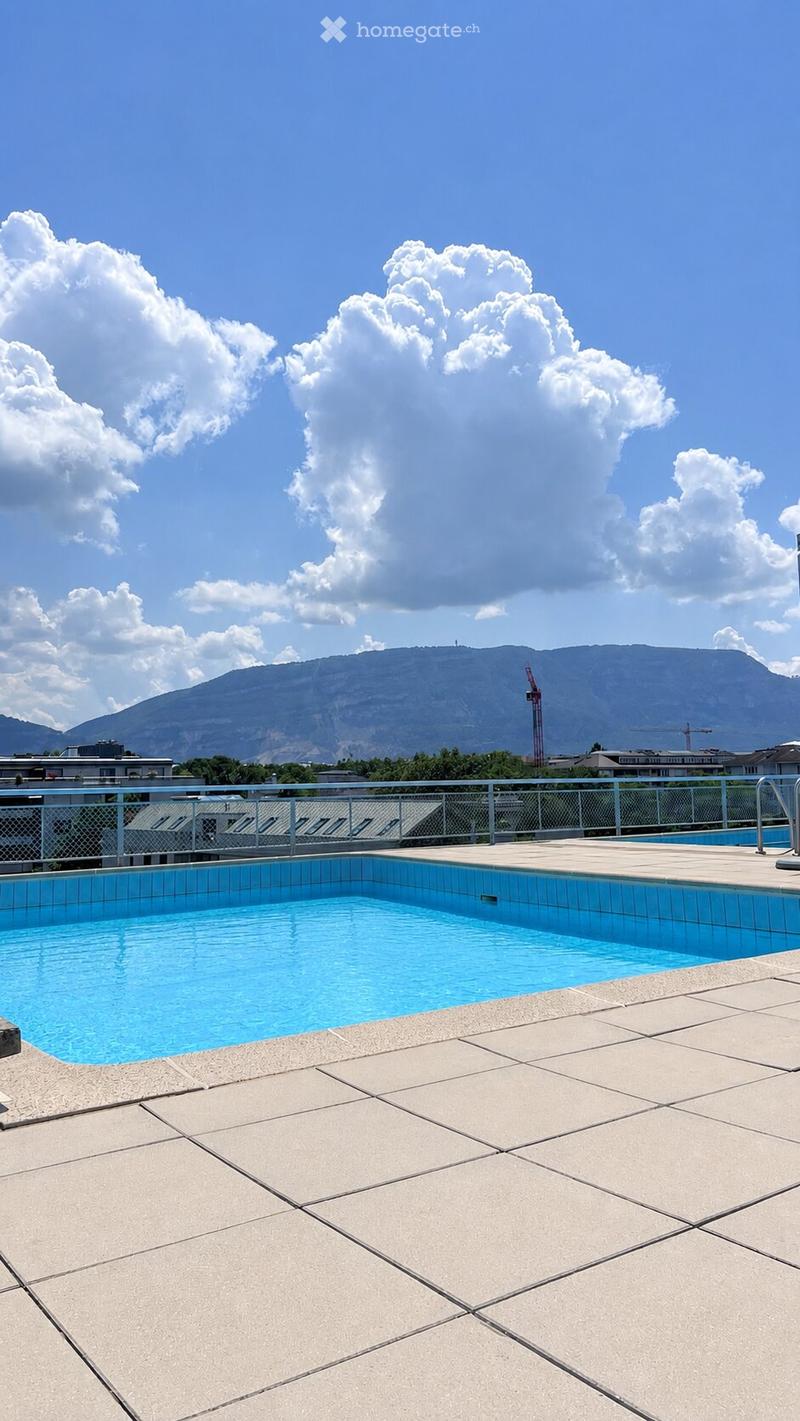 Roof terrace with a swimming pool, surrounded by a metal fence, overlooking a mountainous landscape under a partly cloudy sky.