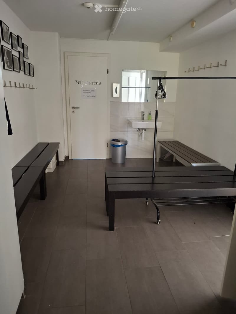 Modern bathroom with dark wooden benches, a white sink, a large mirror, and a trash can. The walls are tiled, and there is a door labeled "WC/Dusche" (toilet/shower).