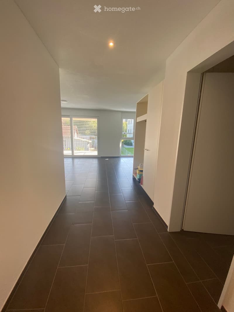 Empty hallway with dark tiled flooring, white walls, and recessed lighting. A small shelf with items is visible on the right side, and a sliding glass door leads to an outdoor area.