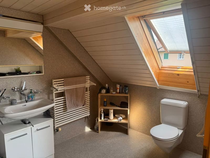 Cozy attic bathroom with wooden slanted ceilings, skylight window, white sink, toilet, towel rack, and small shelf with toiletries.