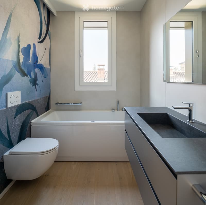 Modern bathroom with a bathtub, toilet, sink, and mirror. Features a large window with a view of a rooftop, and a decorative wall mural with blue floral designs.