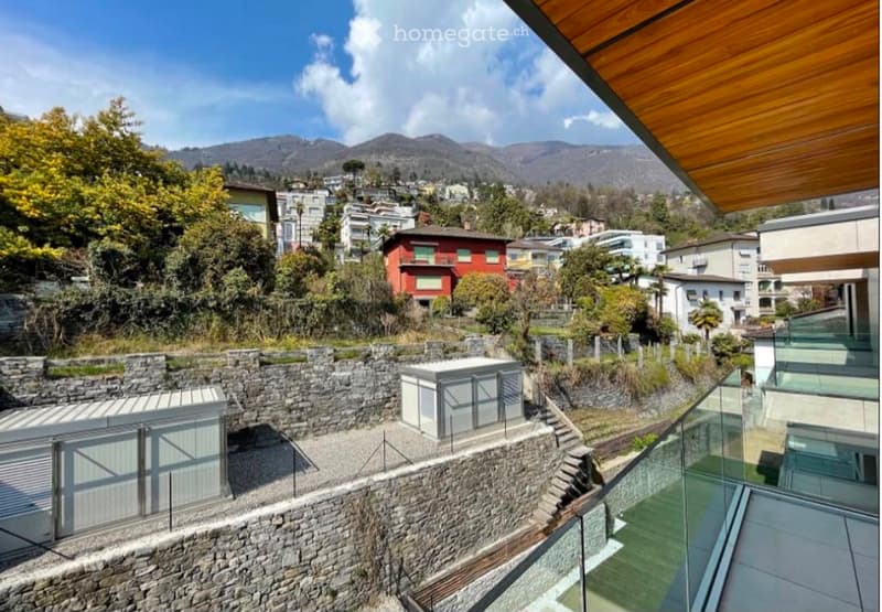 Balcony with a scenic view of a town and mountains, featuring stone walls, modern glass railings, and a small utility shed.