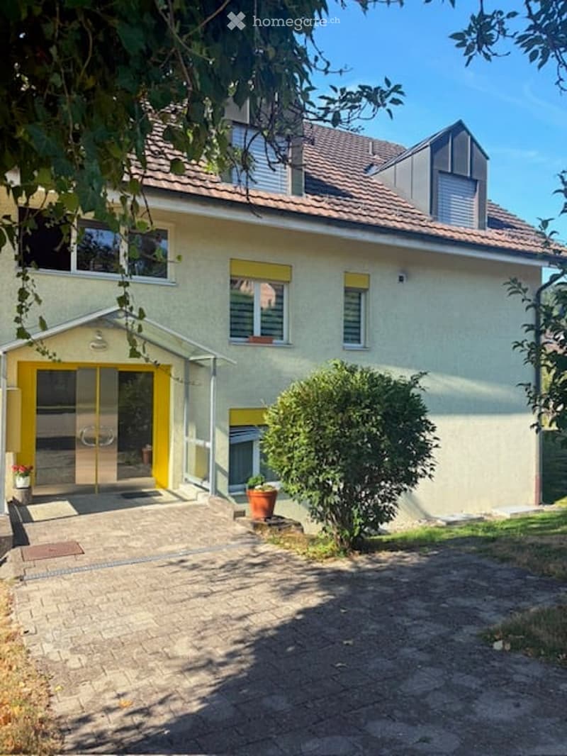 Two-story house with a beige facade, red-tiled roof, and yellow-framed entrance. The entrance features glass doors and a small covered porch area. Potted plants are placed near the entrance, and the house is surrounded by a paved driveway.