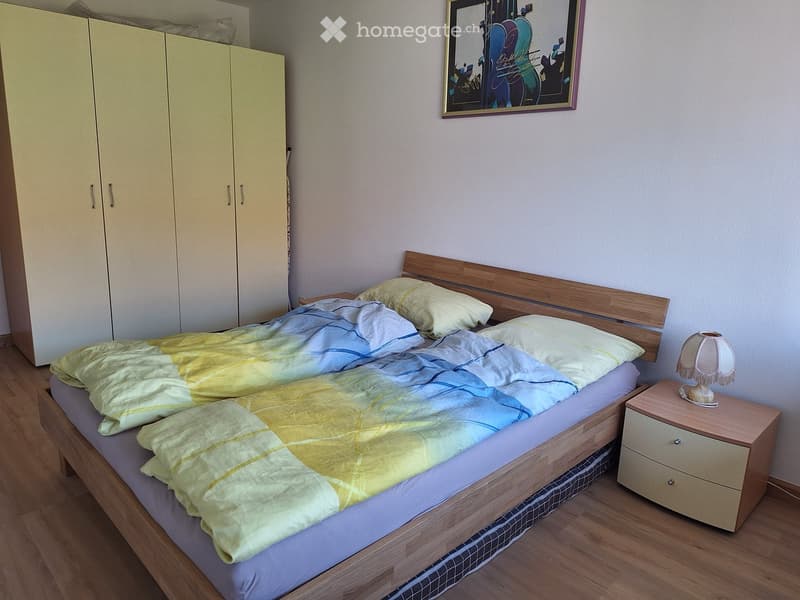 A well-lit bedroom with a wooden bed, a nightstand with a lamp, a wardrobe, and a colorful bedspread.