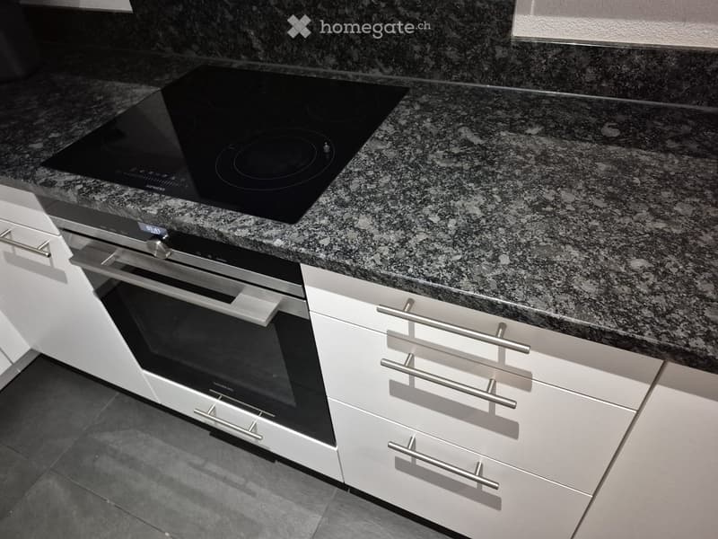 Modern kitchen with granite countertop, induction cooktop, built-in oven, and white cabinets with silver handles.