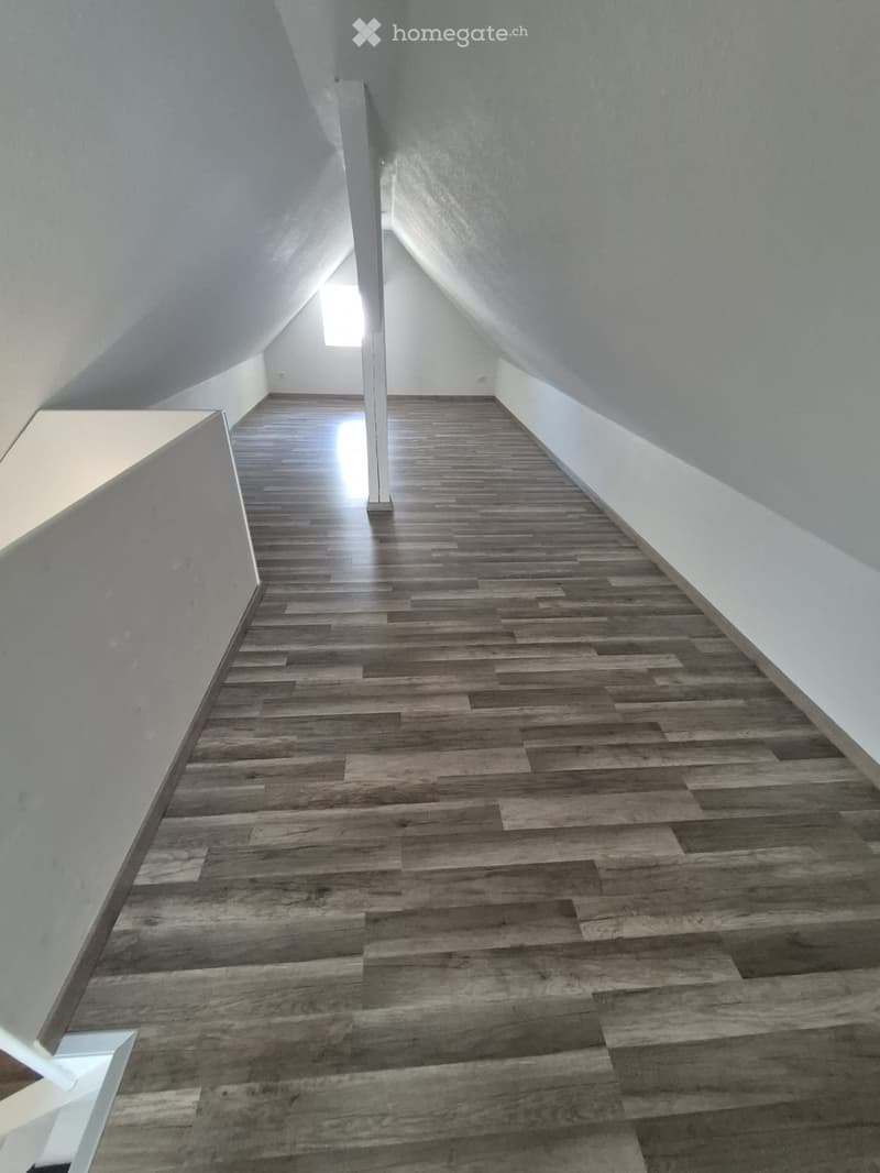 A narrow, slanted hallway with wooden flooring and white walls, featuring a small window at the far end.