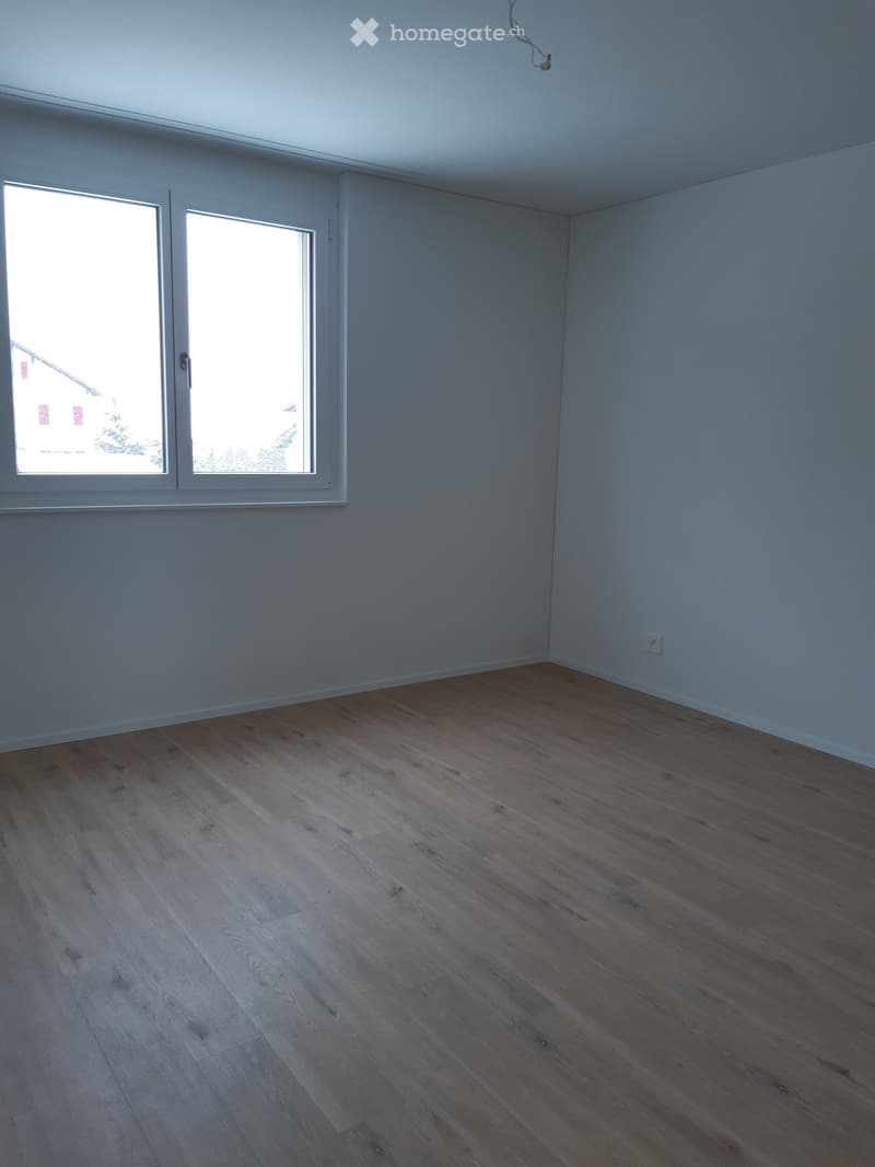 Empty room with light grey walls, wooden flooring, and large windows allowing natural light to enter.