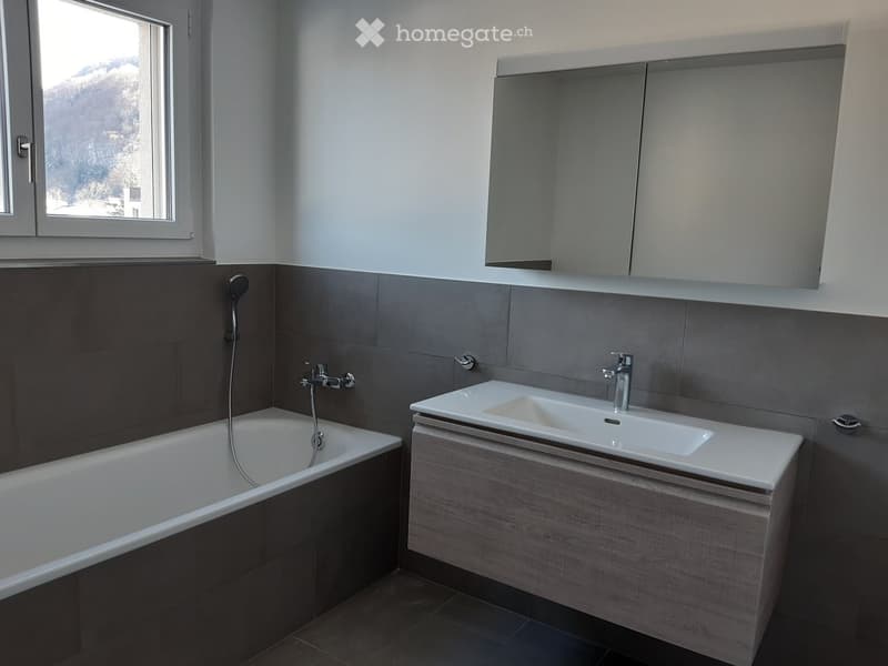 Modern bathroom with a bathtub, sink, mirror, and grey tiled walls.
