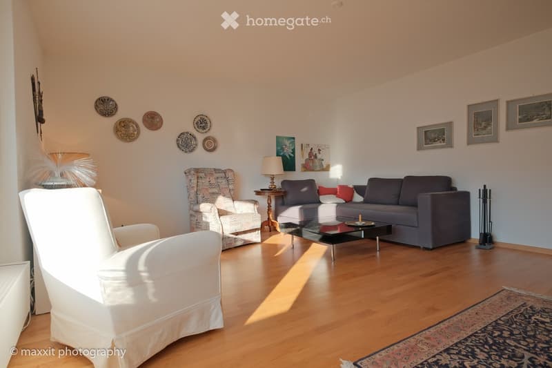 Living Room. Bright living room with wooden flooring, white walls, and large windows allowing natural light. Features a grey sectional sofa, white armchair, coffee table, table lamp, and decorative plates on the wall.