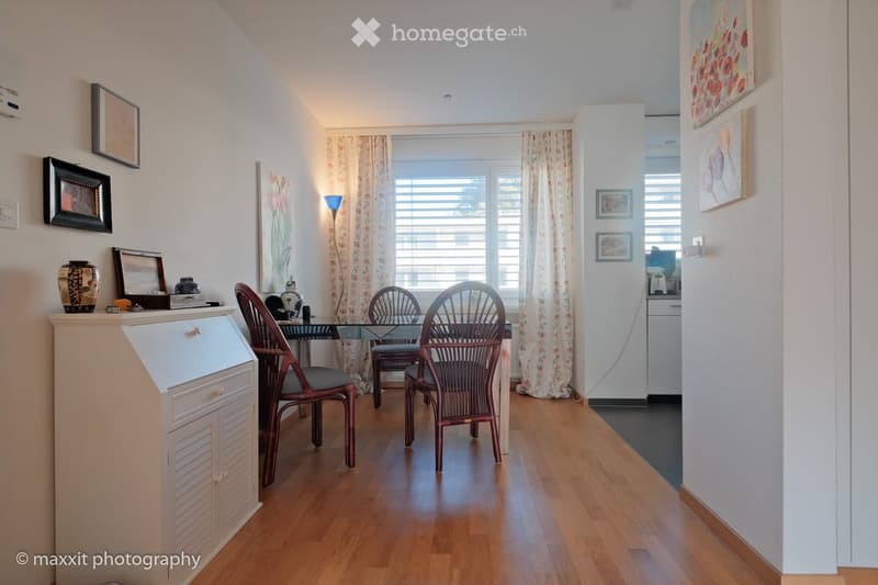 Furnished Room. A well-lit room with wooden flooring, a glass table surrounded by chairs, a small cabinet, and decorative items on top, along with framed artwork on the walls.