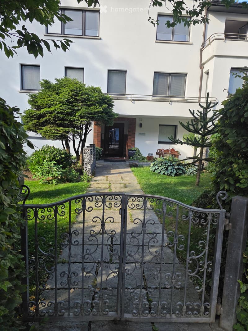 A white multi-story building with multiple windows, a small garden area, and a decorative metal gate leading to the entrance.