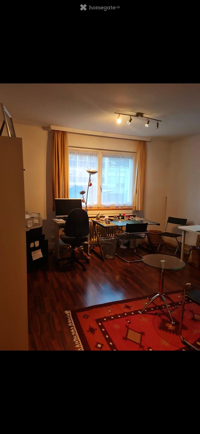 Well-lit room with wooden flooring, large window with curtains, desk with chair, screen, printer, and a round glass table with chairs.