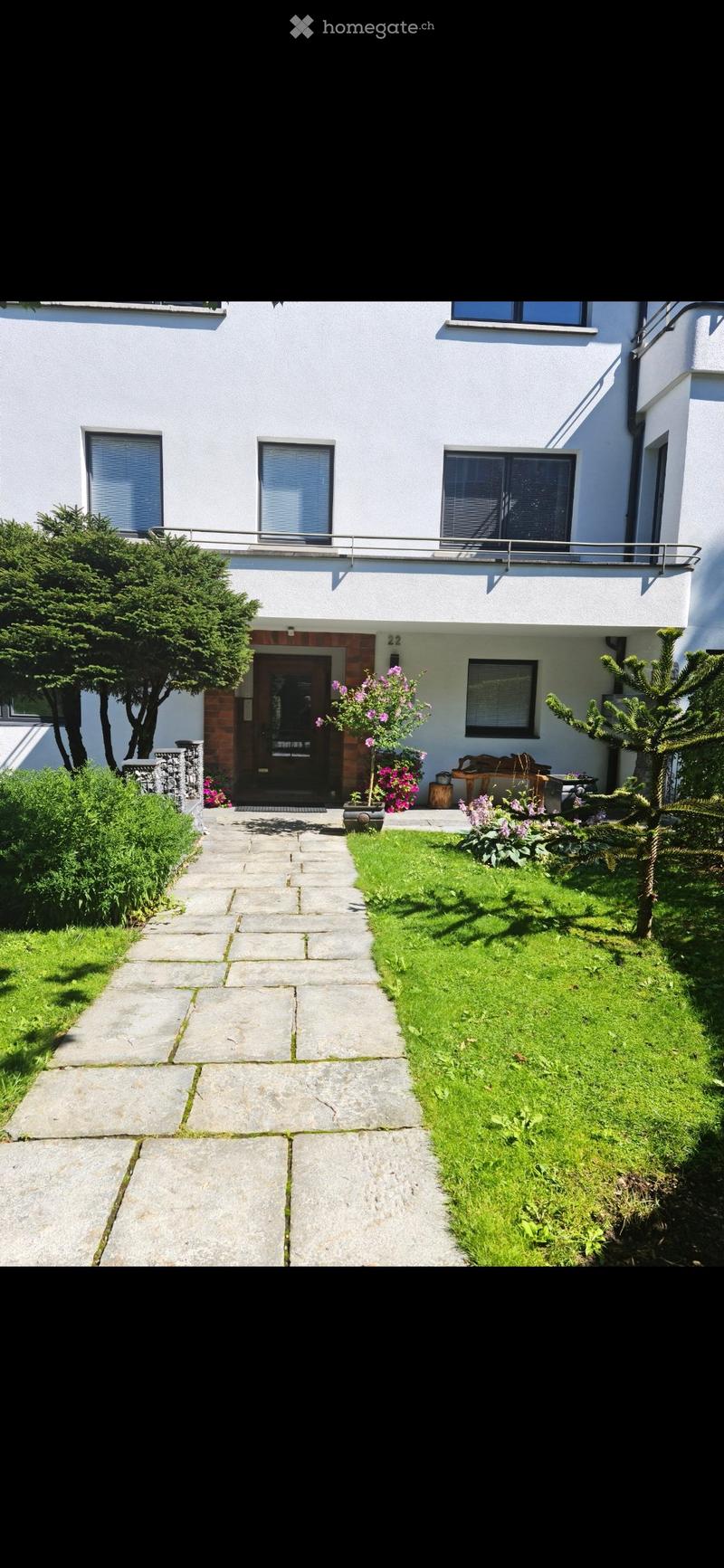 Front garden with a stone pathway leading to a modern white house entrance, surrounded by green grass, shrubs, and flowering plants.