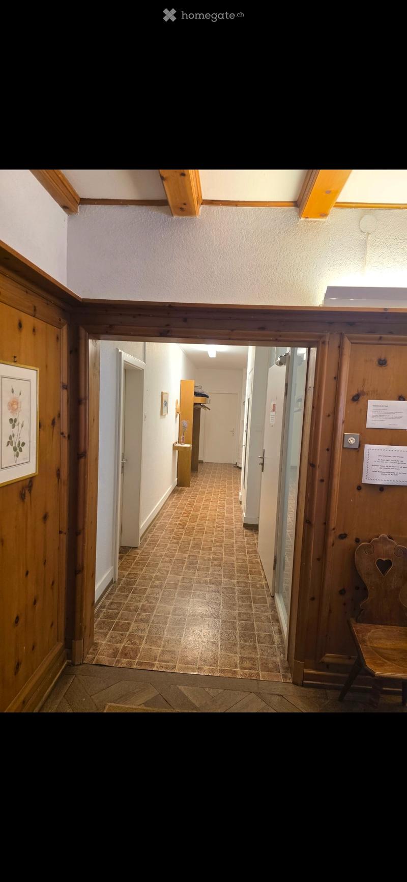 A long, narrow hallway with wooden paneling on the walls, tiled flooring, and several doors leading to different rooms. There is a small wooden bench on the right side and a framed picture on the left wall.