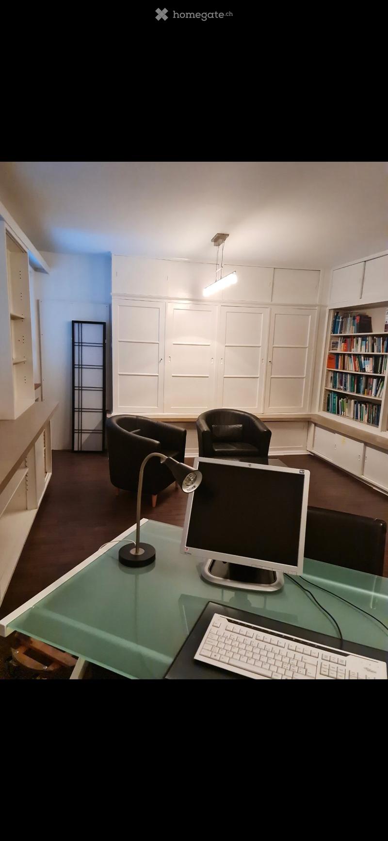 Office space with a glass desk, computer setup, two armchairs, a table lamp, and built-in white cabinetry with a bookshelf.