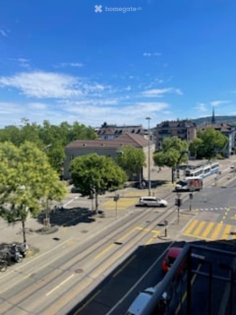 A view of a city street from a balcony or terrace, showing a road with traffic, buildings, trees, and a clear blue sky.