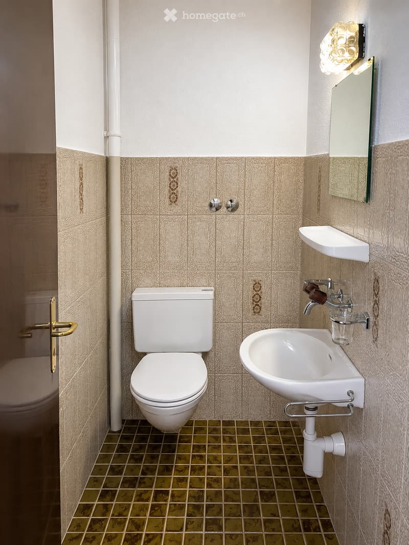 A compact bathroom with beige patterned tiles, a white toilet, a wall-mounted sink with a chrome faucet, and a mirrored cabinet with a built-in light fixture.