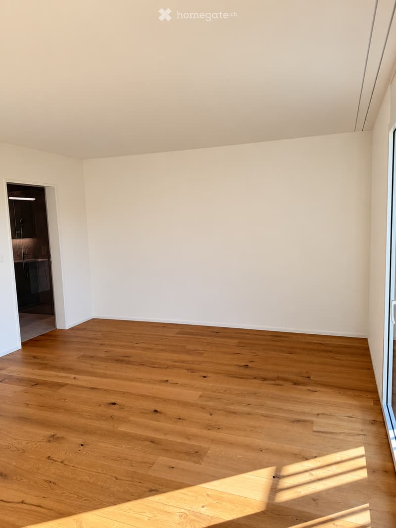 Empty room with light-colored walls and polished wooden flooring, featuring a doorway leading to another room and a large window allowing natural light to enter.