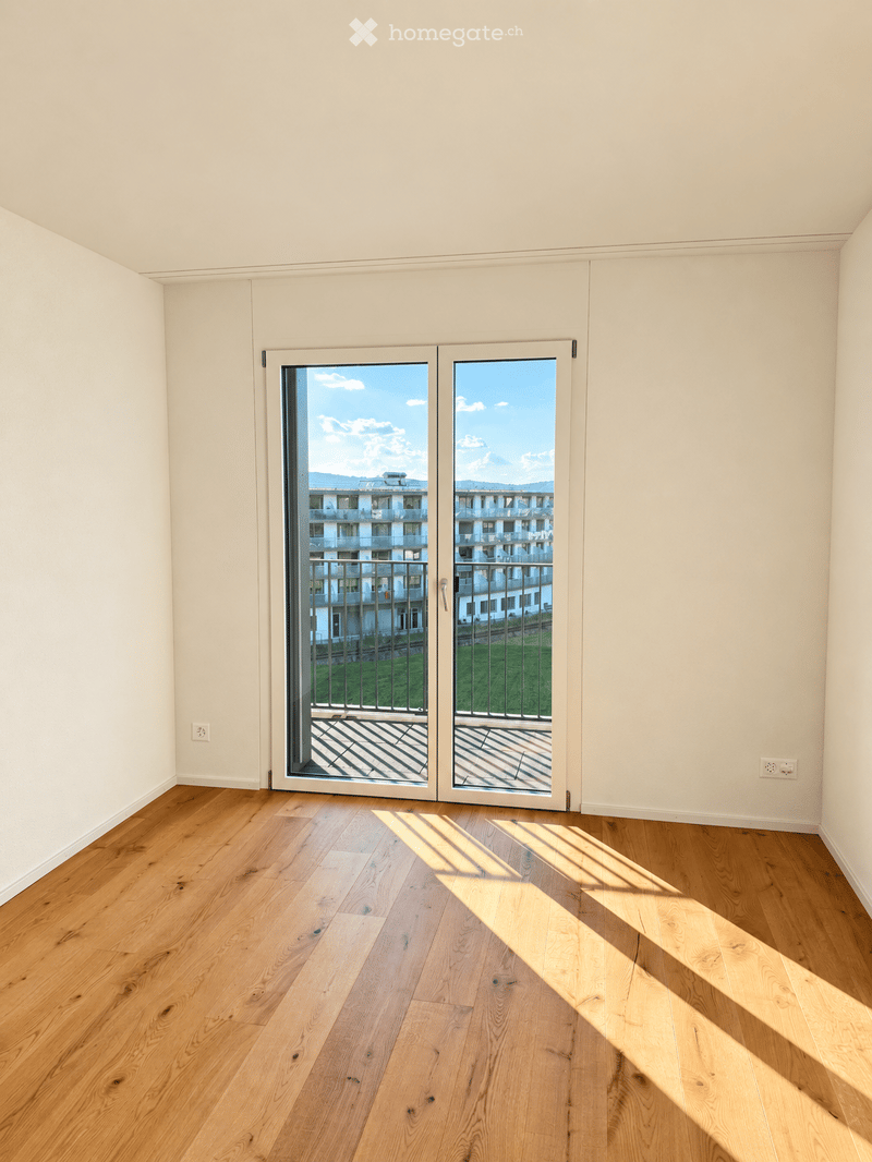 Empty room with light-colored walls, wooden flooring, and large sliding glass doors leading to a balcony.