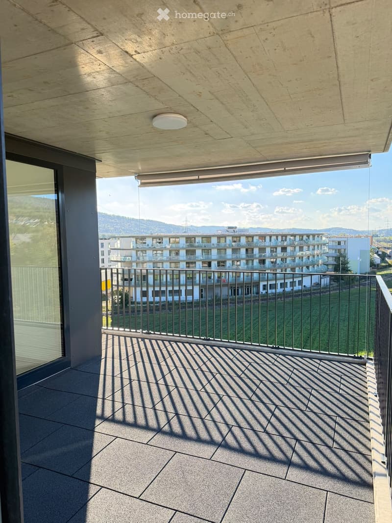 Modern balcony with concrete floor, metal railing, and large windows offering a view of the surrounding area and mountains.