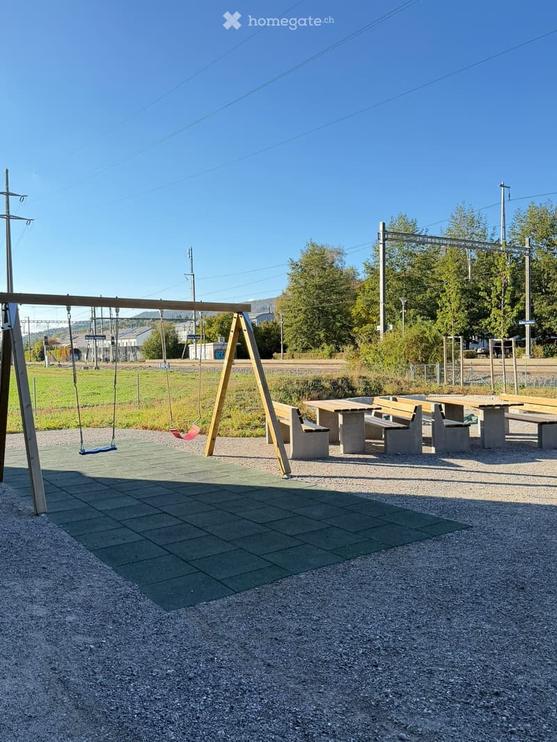 Outdoor park area with swings, benches, and tables on a gravel surface, surrounded by greenery and a clear blue sky.