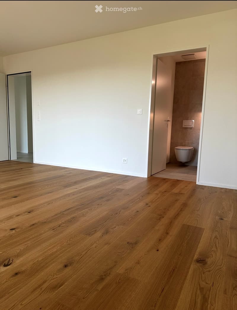 Empty room with light wooden flooring and white walls, featuring an open doorway to a small bathroom with a toilet.