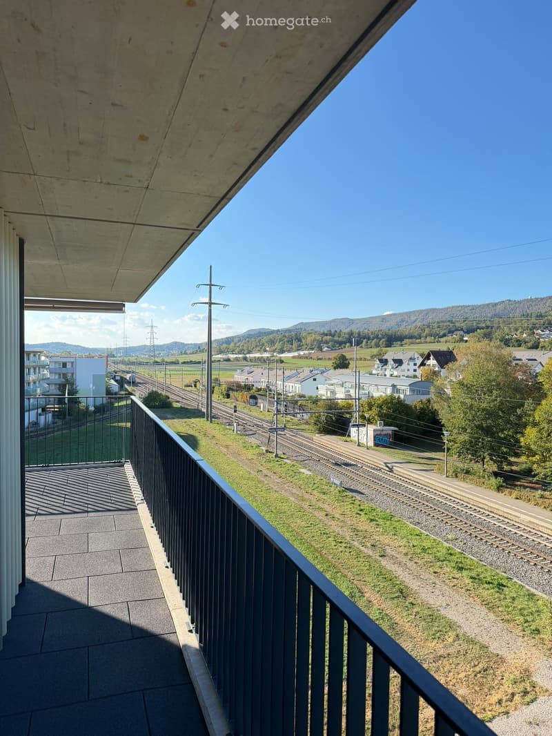 Balcony with a metal railing overlooking a railway track and surrounding landscape with houses and hills.