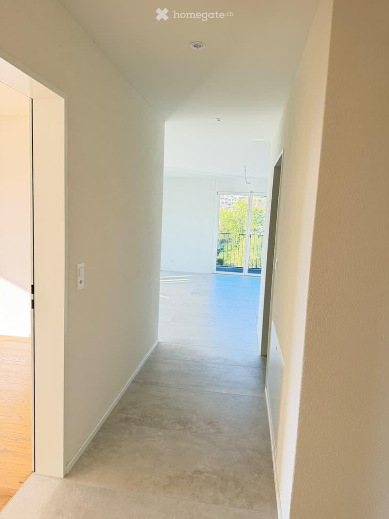Empty hallway with light-colored walls, tiled floor, and recessed lighting. An open door leads to a room with wooden flooring, and a sliding glass door opens to a balcony with outdoor view.