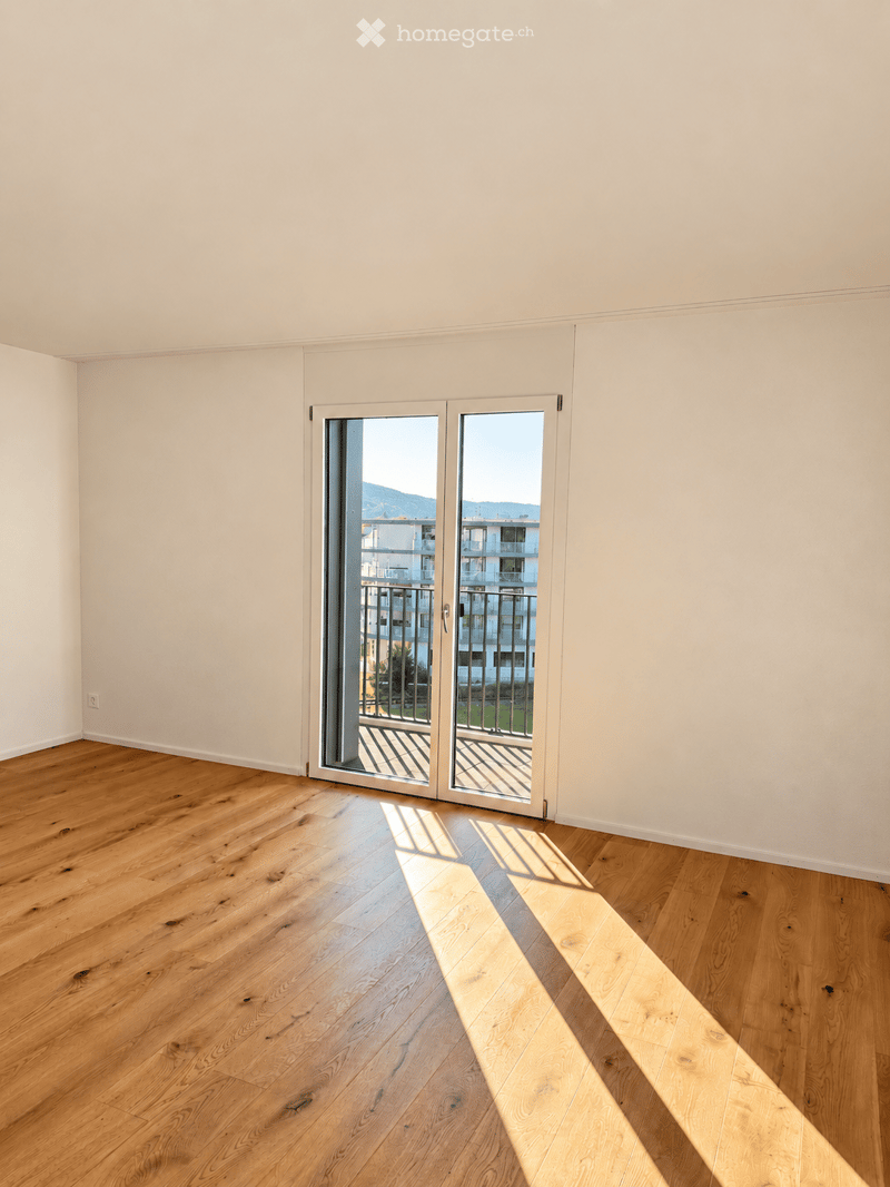 Empty room with light wooden flooring and white walls, featuring a large sliding glass door leading to a balcony with a view of buildings and mountains.