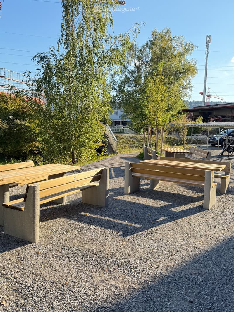 Outdoor seating area with two wooden benches and tables on a gravel surface, surrounded by trees and greenery.