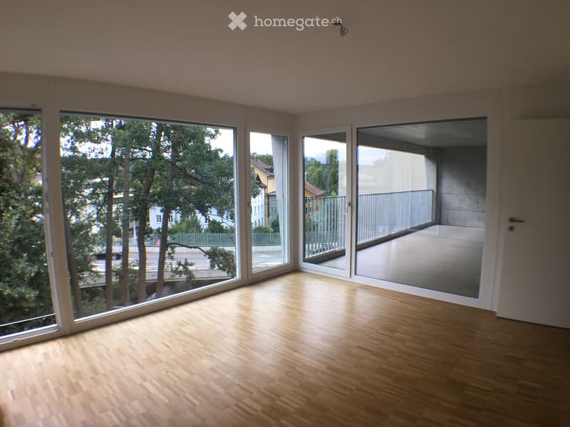 Empty room with wooden flooring and large windows offering a view of trees and a street. The room has a sliding glass door leading to a balcony area.